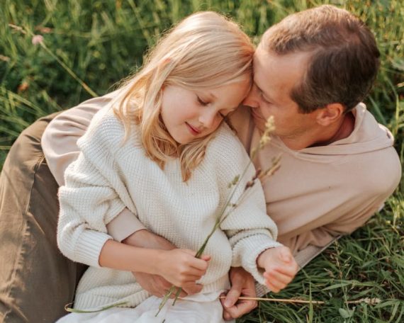 A father and daughter share a loving moment in a grassy field, enjoying nature together.